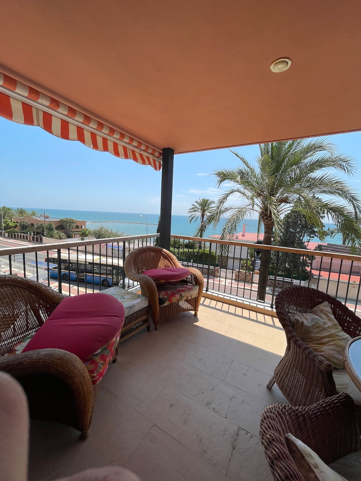 A balcony overlooking the sea is furnished with wicker seating and cushions. Sunlight illuminates the space, creating a comfortable outdoor area. Palm trees are visible in the background, along with a view of the beachfront and distant surroundings.