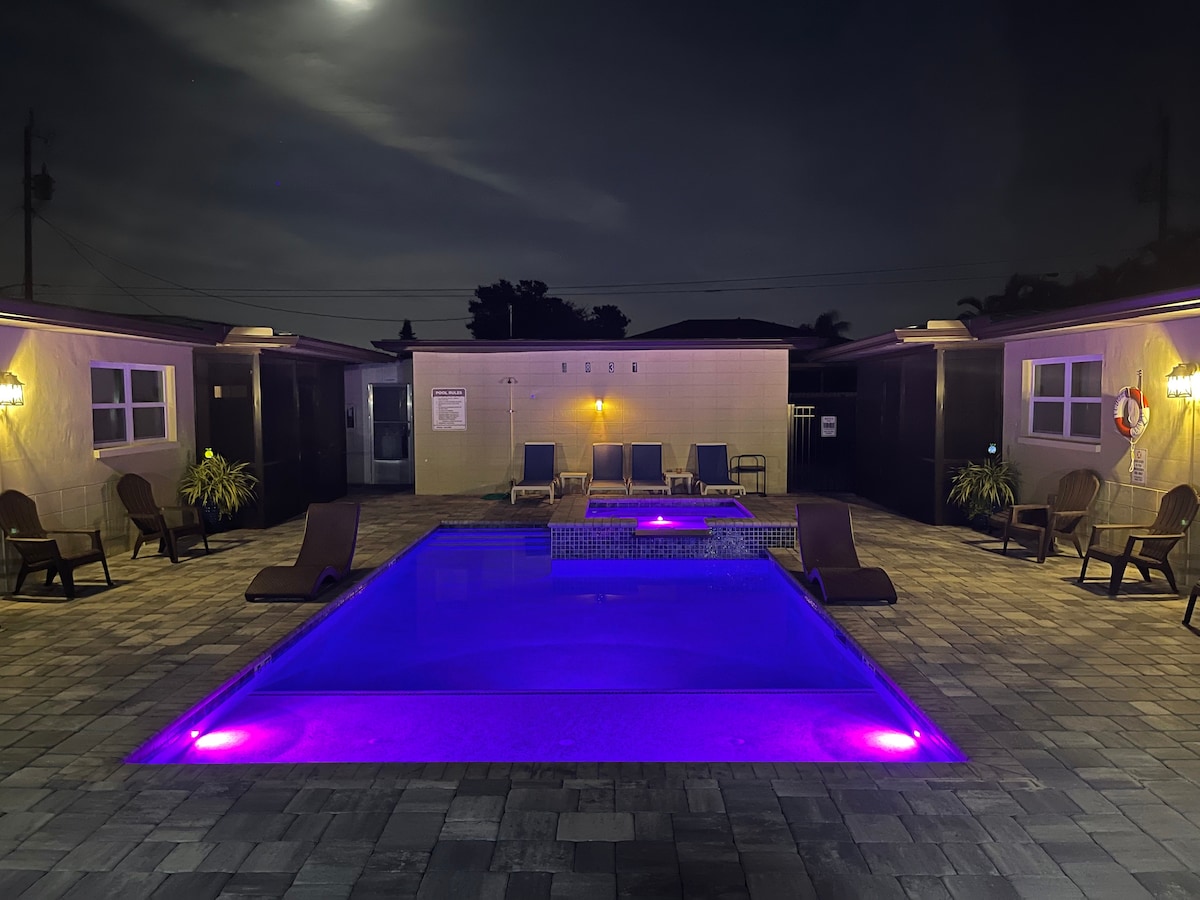 The illuminated heated pool and spa are surrounded by a spacious deck, featuring lounge chairs for relaxation. Soft purple lighting enhances the tranquil atmosphere, while the pool area is bordered by the exterior of the apartment complex, emphasizing privacy and comfort.