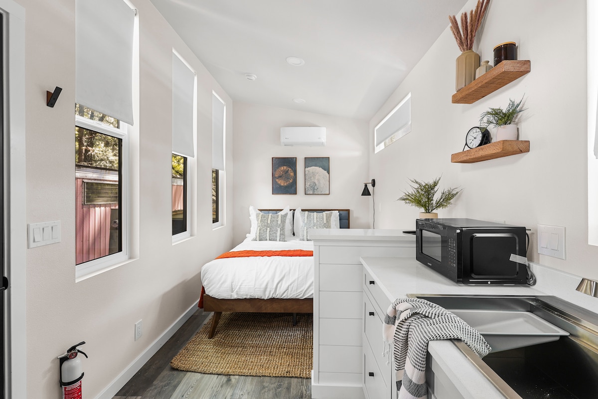 A studio interior features a queen-sized bed with crisp white linens and an orange accent blanket. Wall shelves display decorative items. A microwave is positioned on the countertop near a sink. Multiple windows provide natural light, and an area rug adds warmth to the wooden floor.