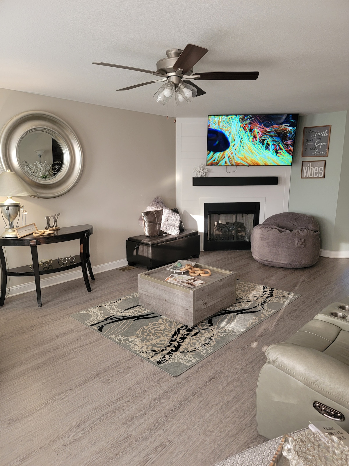 The living room is designed for comfort, featuring a large wall-mounted TV, a cozy fireplace, and a soft bean bag. A round mirror reflects light above a small table next to an elegant side table. A decorative area rug adds visual interest beneath a central coffee table.