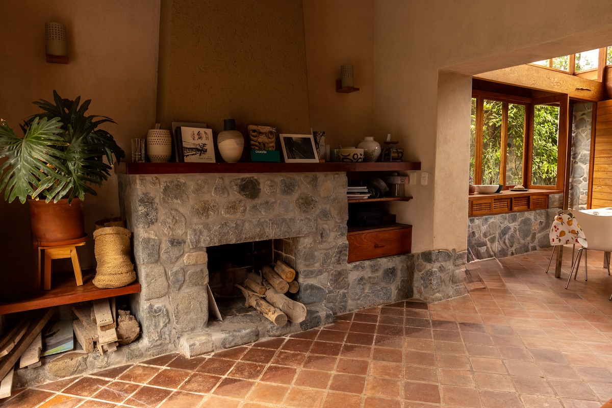 An interior space features a stone fireplace with neatly stacked logs in front. A wooden shelf displays decorative items and books. Sunlight filters through large windows, illuminating the warm tile floor and connecting to a bright dining area.