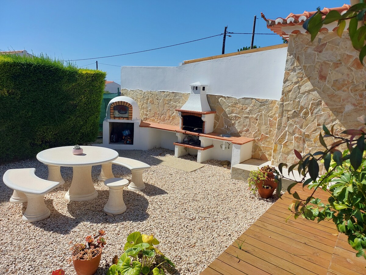 An outdoor area features a rustic stone barbecue station alongside a circular table surrounded by four matching benches. The space is bordered by lush greenery and gravel, providing a cozy setting for outdoor meals and gatherings.