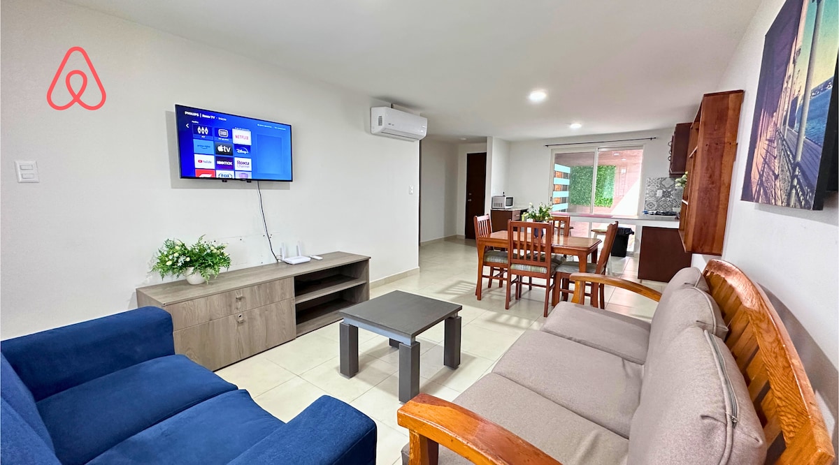 The living area features a blue sofa and a beige couch, accompanied by a coffee table and a wooden dining table with four chairs. A mounted television is visible on the wall, and natural light enters through nearby windows.