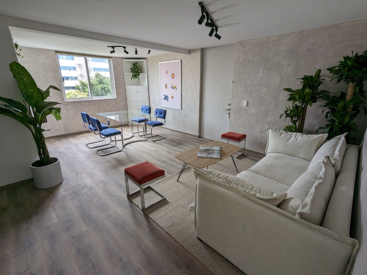 A bright living space features a comfortable sofa, modern coffee table, and colorful chairs. Lush plants are positioned in the corners, contributing to a vibrant atmosphere. Large windows allow natural light to fill the area, enhancing the airy feel of the room.