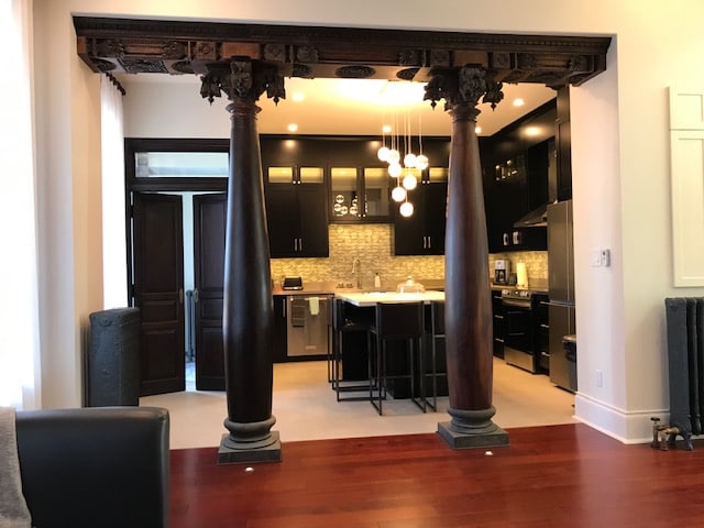 The image captures an open-concept kitchen, framed by two intricately carved wood columns. Modern cabinetry and stainless steel appliances are visible, along with a large island surrounded by high stools. The lighting is softened by stylish pendant fixtures, creating an inviting cooking space.