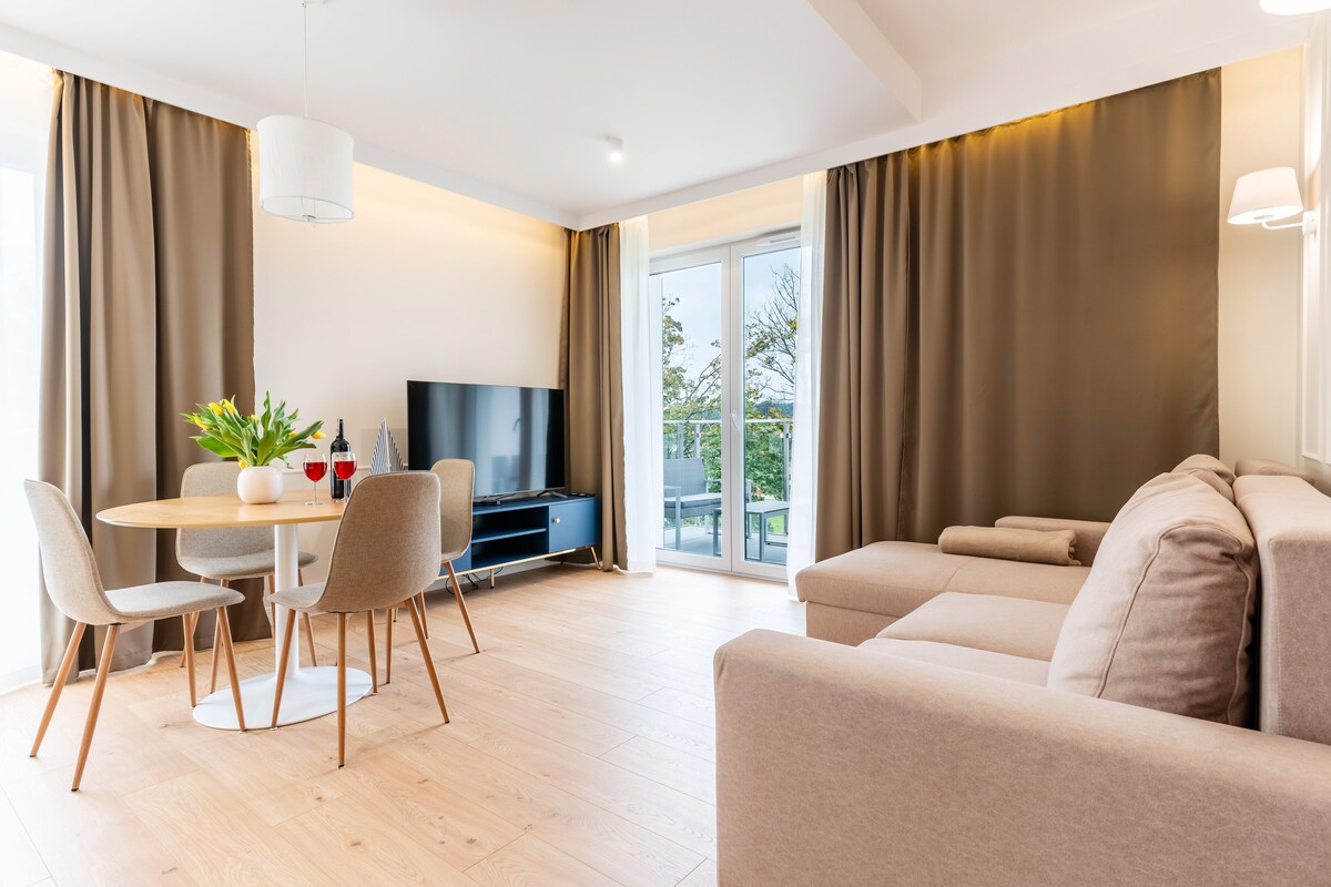 A bright and airy living area features a comfortable beige sofa facing a sleek television. A round dining table with light-colored chairs is set near a large glass door that opens onto a balcony, allowing natural light to enhance the space.