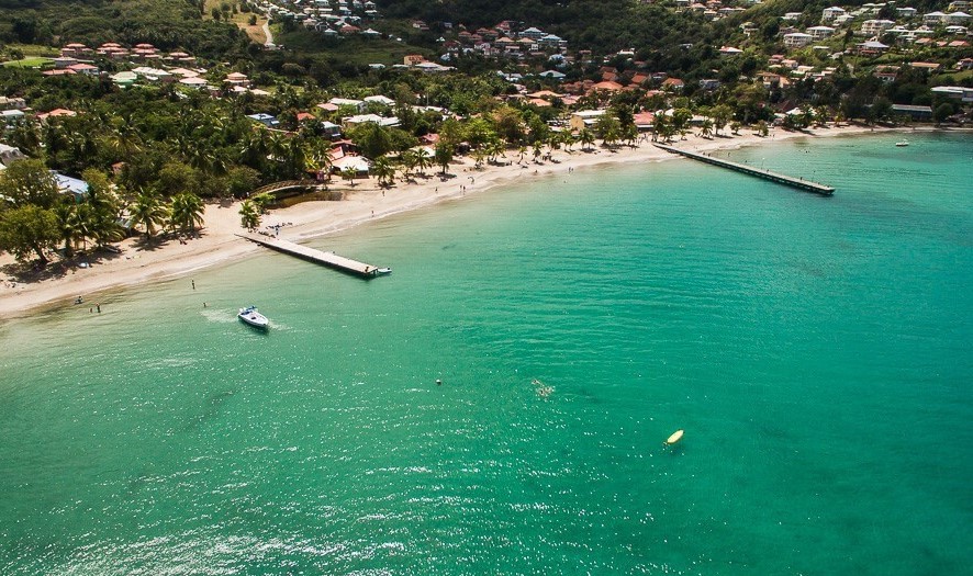 Aerial view of a serene beach with clear turquoise waters gently lapping at the shore. Several people can be seen enjoying the sandy beach and a few boats are anchored nearby. Lush greenery and residential areas frame the coastline, enhancing the natural setting.