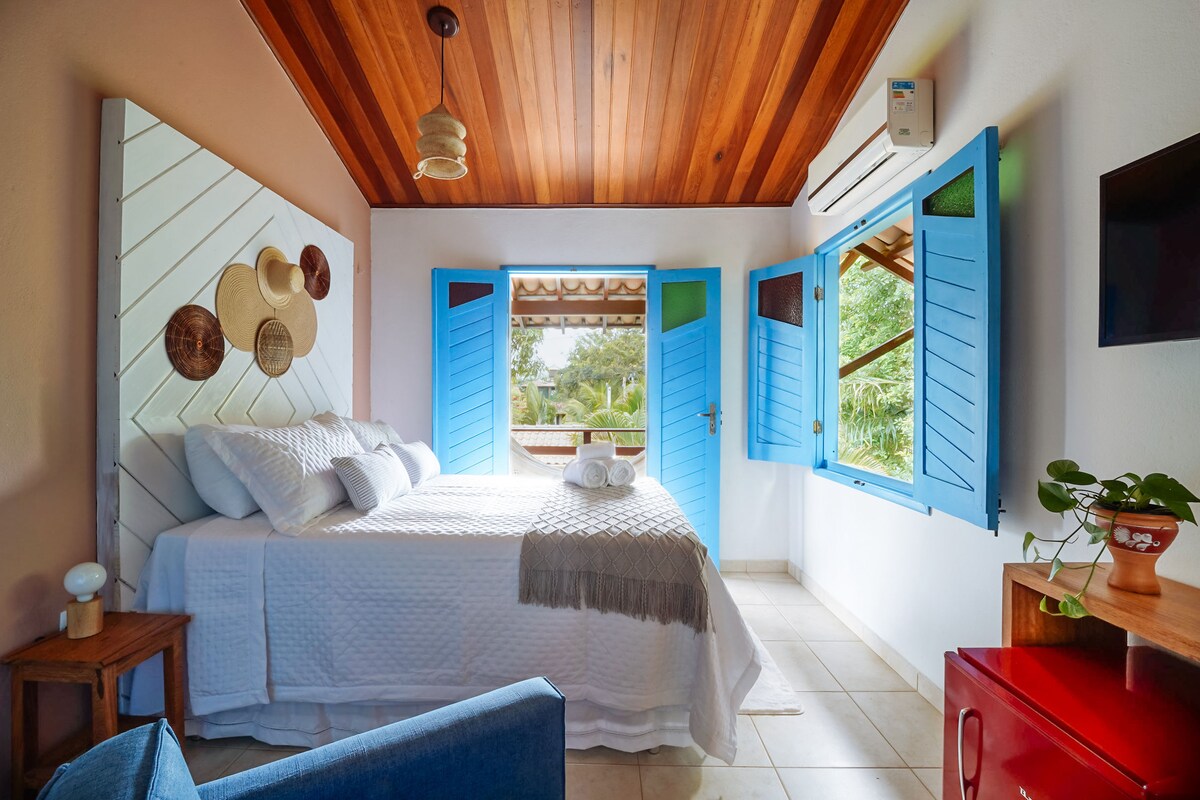 A comfortable suite features a spacious bed with soft bedding, framed by bright blue shutters. Natural light floods the room, highlighting wooden ceiling beams. A small plant rests on a side table, while a cozy seating area invites relaxation.