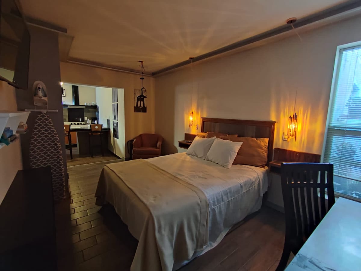 A cozy bedroom features a spacious bed dressed in neutral bedding. Warm lighting emanates from wall sconces, creating a relaxed ambiance. A comfortable chair is positioned nearby, while a kitchenette is visible in the background, enhancing the practicality of the space.