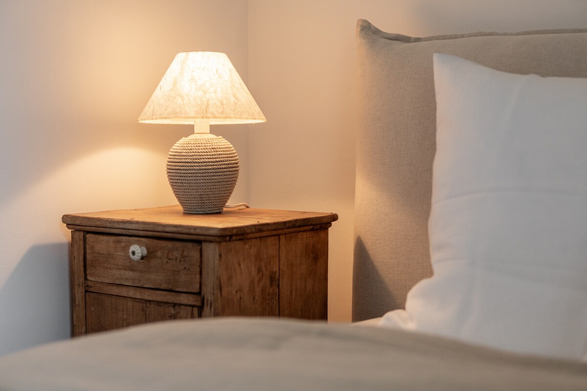 A bedside table made of wood holds a textured lamp with a soft light shade. The warm glow illuminates the cozy sleeping area, which is outlined by a plush headboard and a neatly arranged pillow.