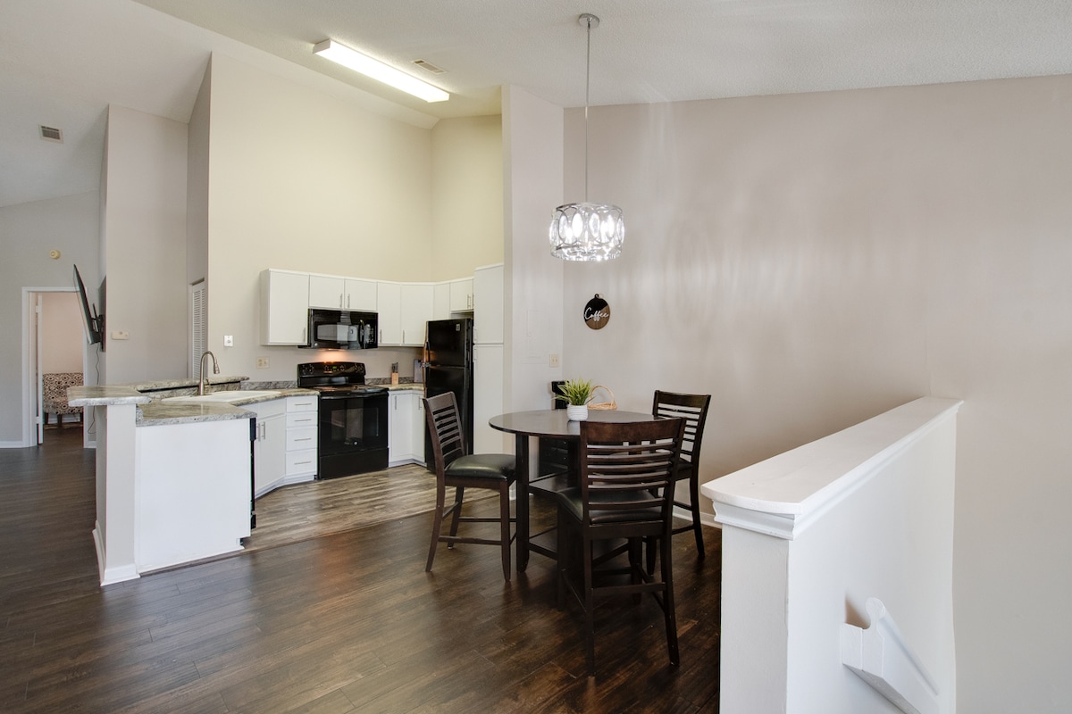 An open dining area is featured, showcasing a round wooden table surrounded by four dark chairs. A modern light fixture hangs above the table. The kitchen, equipped with white cabinets and black appliances, is visible in the background, highlighting a spacious layout.