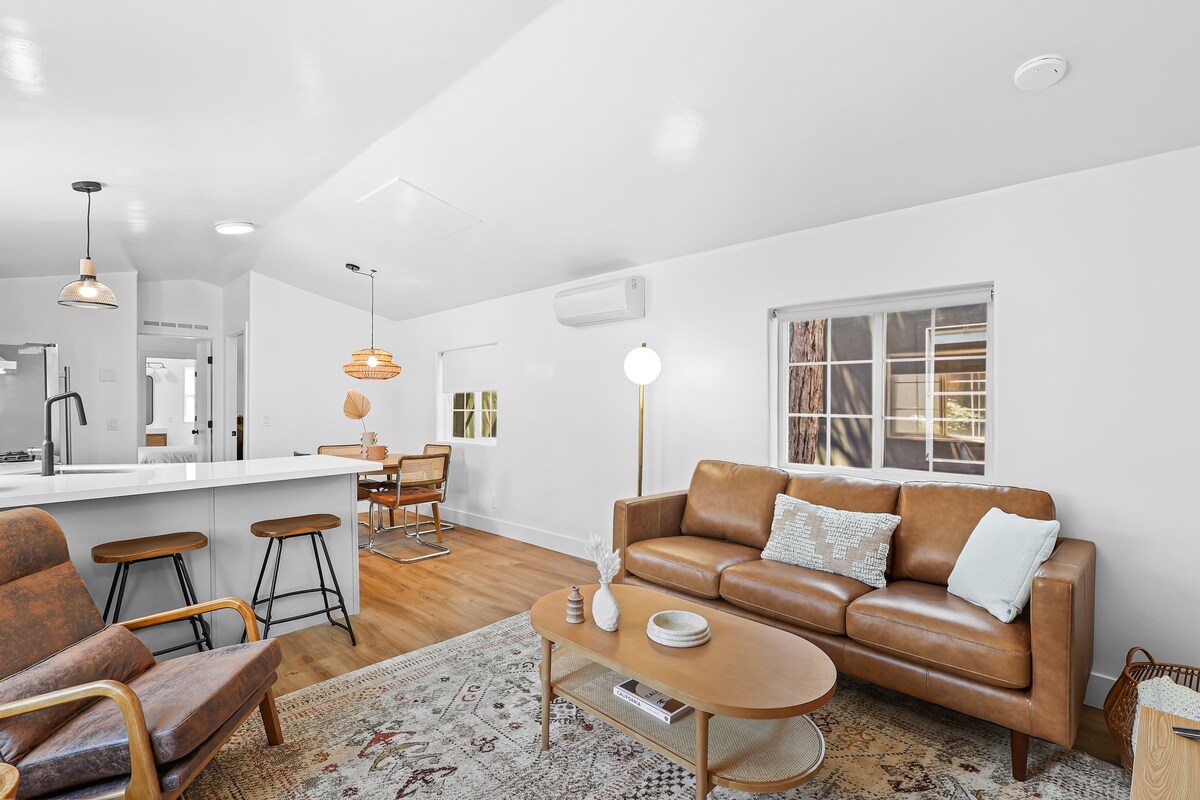 An inviting living space features a comfortable leather sofa, accented with a light blue throw pillow. A round coffee table sits in front, surrounded by wooden chairs. Natural light filters through the window, illuminating the clean white walls and smooth wooden flooring.