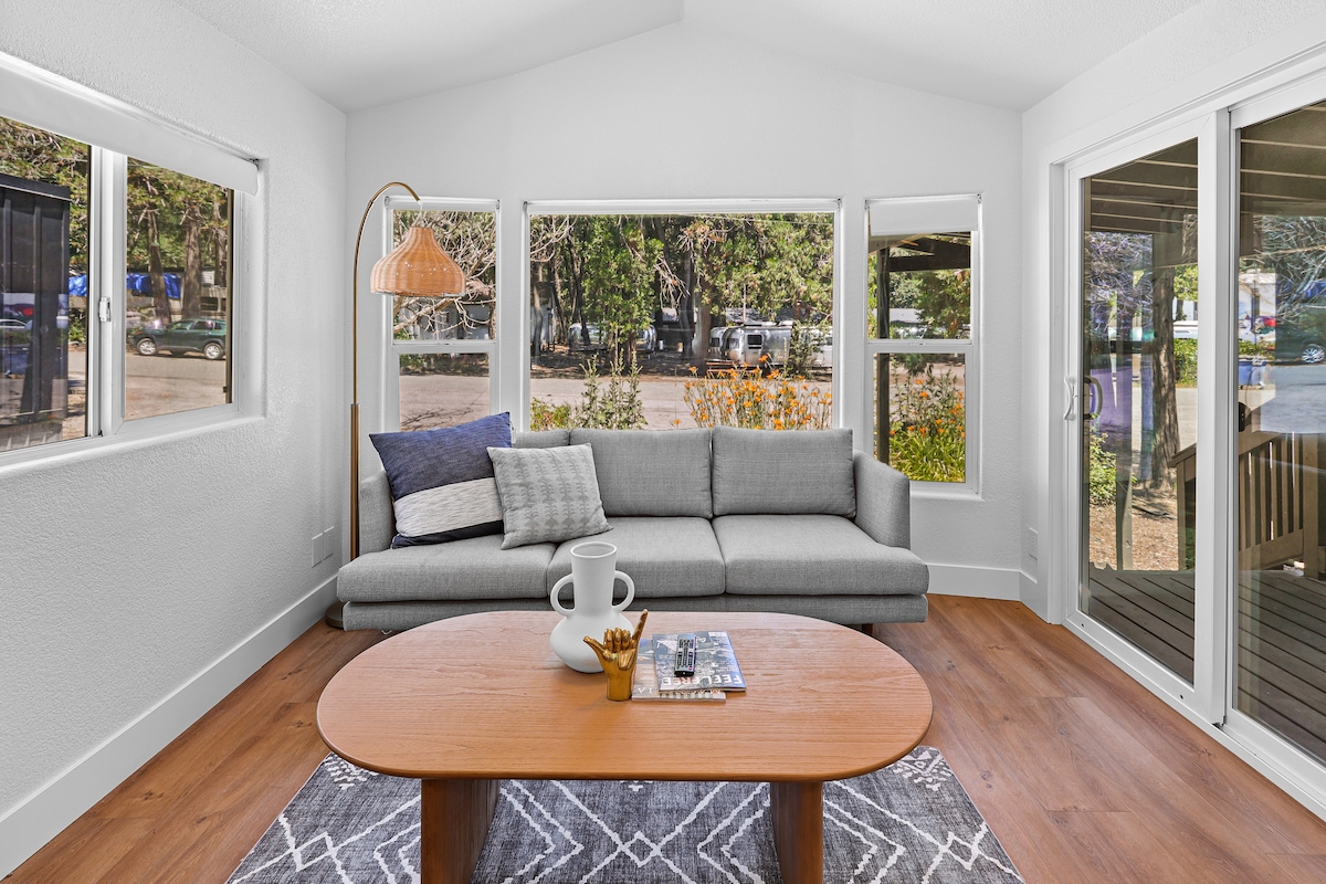 A modern living area is furnished with a light gray sectional sofa and a round wooden coffee table. Large windows allow natural light to fill the space, revealing a view of the outdoor landscape. Decorative elements include a woven lamp and a patterned rug.
