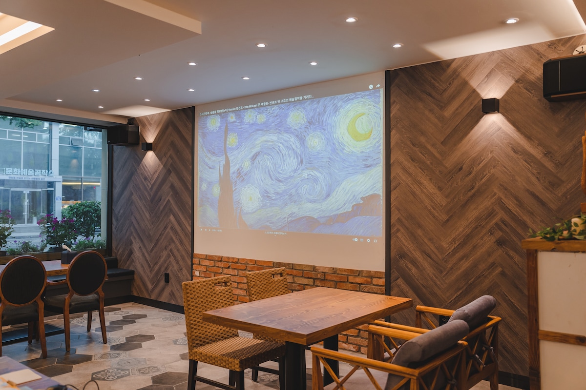 A spacious café area is depicted, featuring a large projection wall displaying a famous starry night scene. The wooden accents and modern lighting create a relaxed ambiance. A wooden table is surrounded by chairs, inviting guests to gather and enjoy the space.