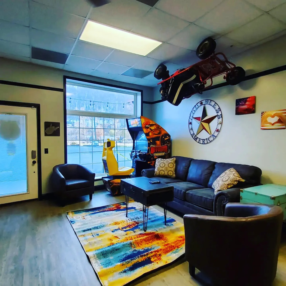 A lively game room is featured, showcasing a large leather sofa opposite a coffee table. An arcade game cabinet and a racing simulator are positioned against the wall. A colorful area rug lies beneath the furniture, and overhead, a small red vehicle is displayed.