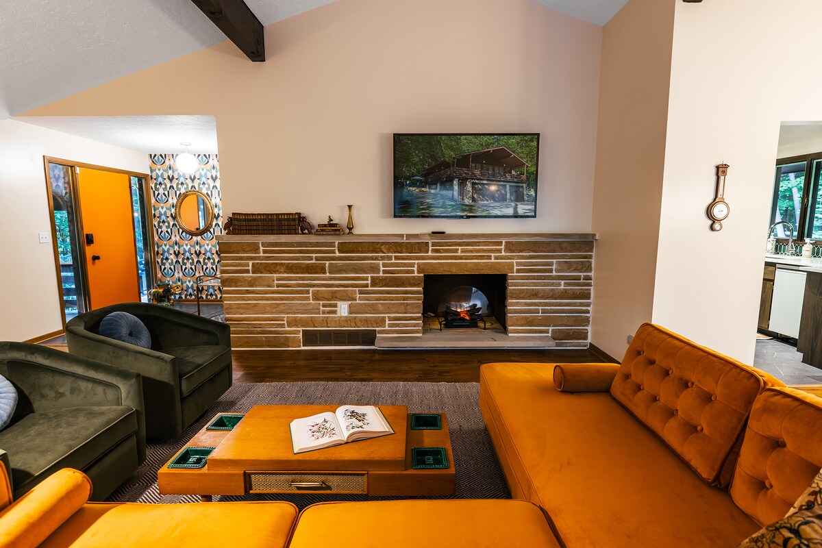 A spacious living area features an orange sectional sofa complemented by two green armchairs. A coffee table with decorative elements sits centrally, while a stone fireplace provides a cozy focus. A large television is mounted on the wall, enhancing the aesthetic.