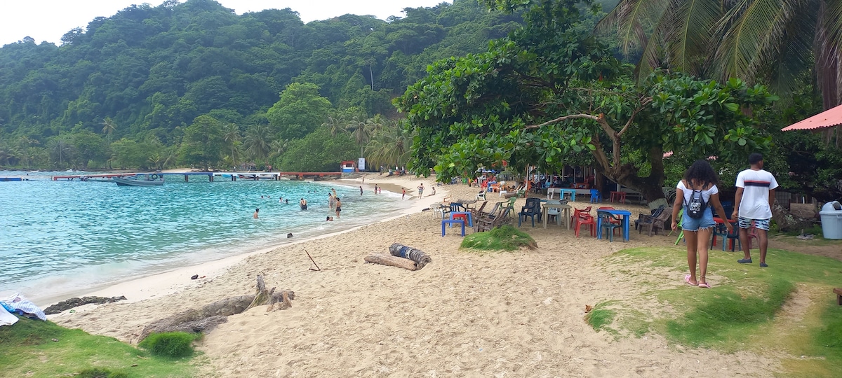 A beach scene is presented, featuring soft sand and clear water. Lush greenery lines the shoreline, while several people enjoy the water. Colorful tables and chairs are arranged under shade trees, creating a relaxed atmosphere. A gentle curve of the beach leads towards visible activity areas.