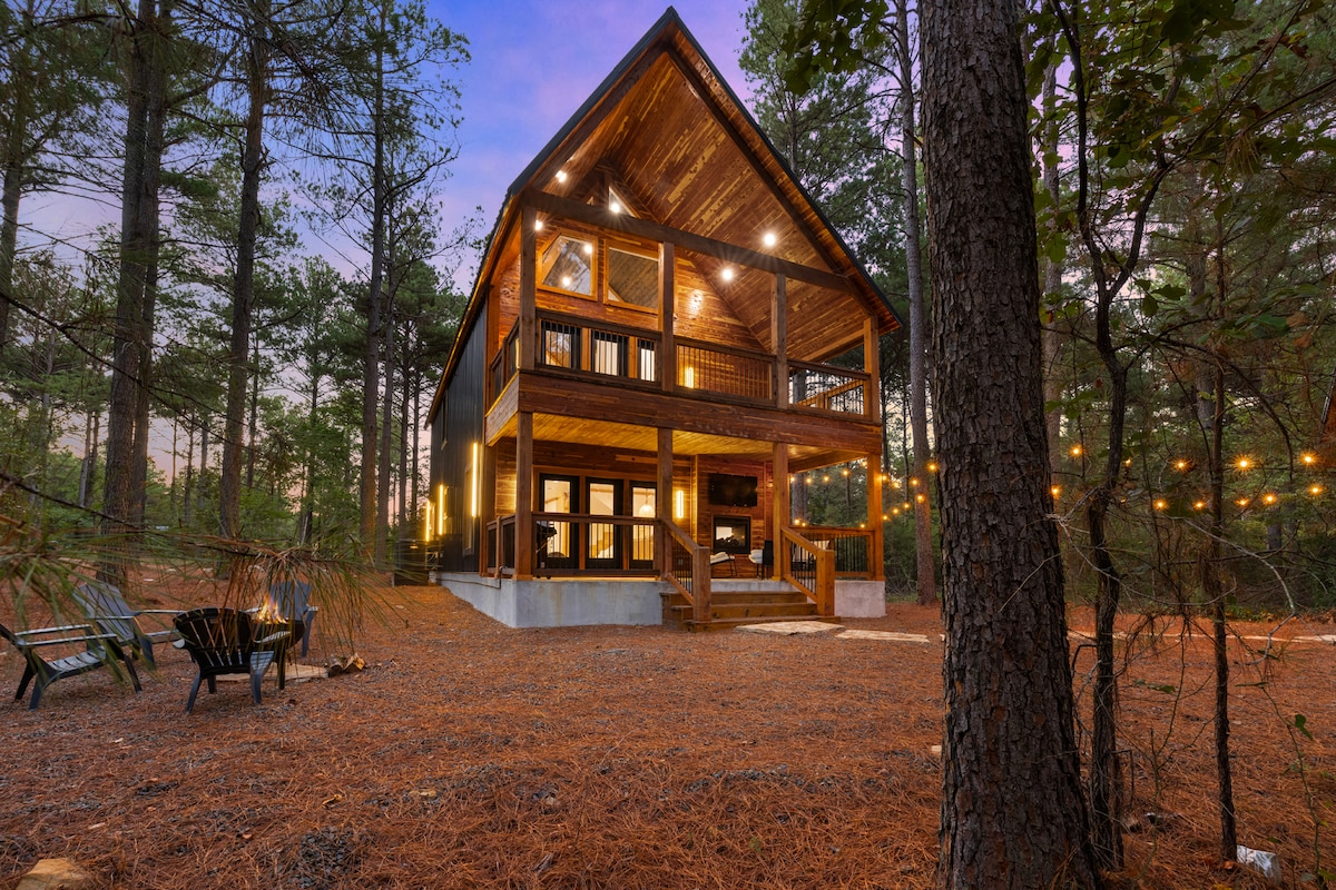 A modern cabin is nestled among tall trees, featuring a wooden exterior and a sloping roof. The deck is illuminated with warm lights, and comfortable seating is arranged in front. A cozy ambiance is created by the surrounding natural setting, enhanced by the soft glow of the evening sky.