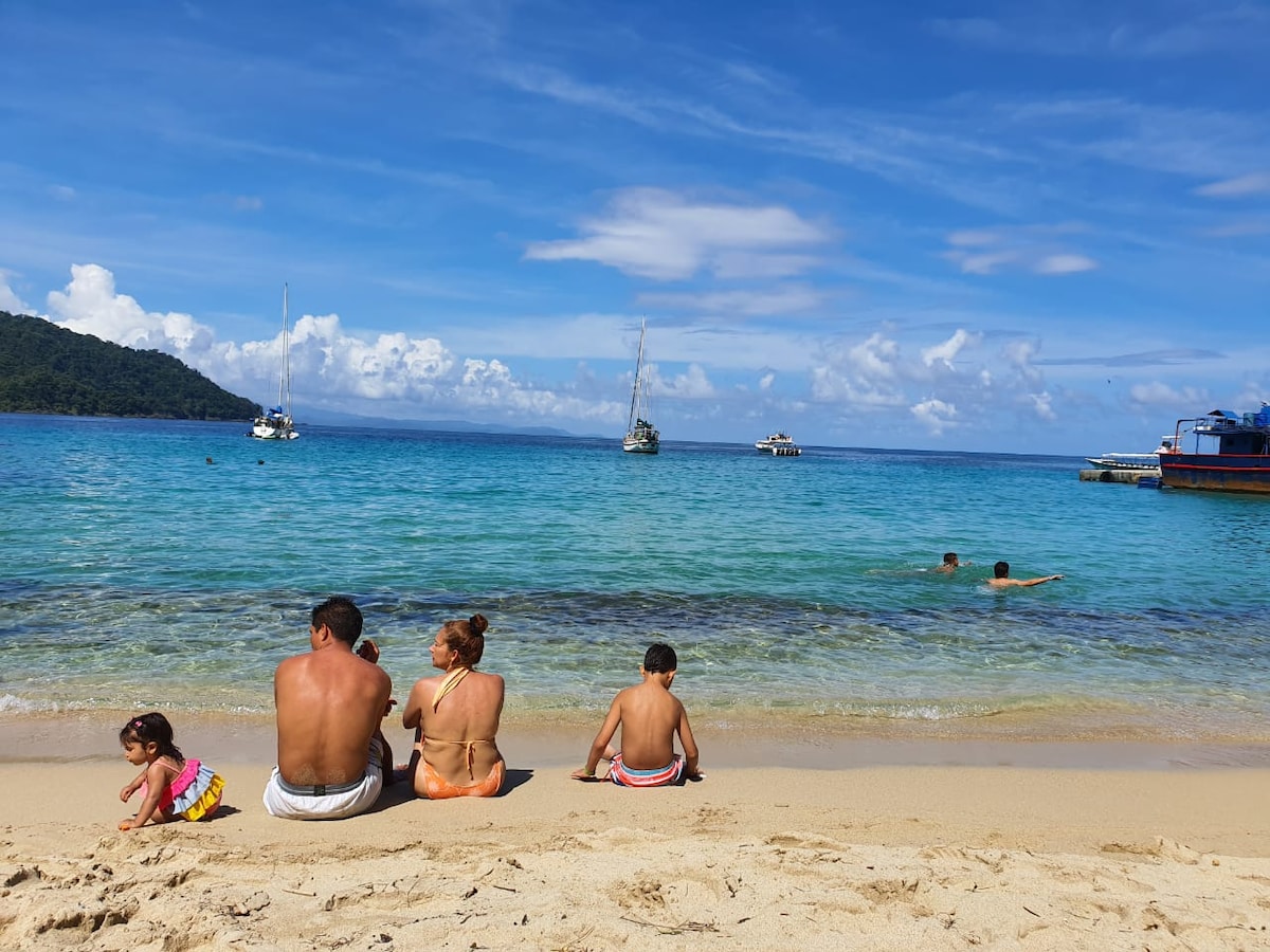 A sandy beach scene is presented, featuring a family sitting together at the water's edge. Calm turquoise waters extend towards a distant horizon, dotted with small boats. Lush green hills rise in the background under a partly cloudy sky.