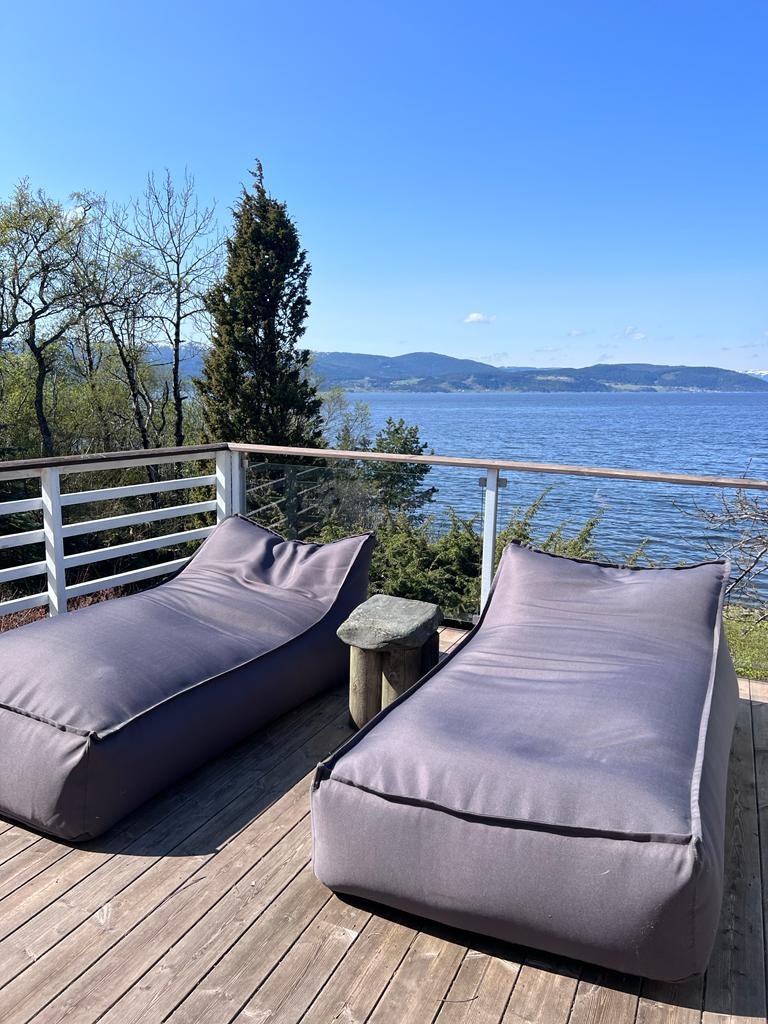 Two spacious outdoor lounge chairs are positioned on a wooden deck, overlooking the tranquil waters of the fjord. The serene landscape features trees and distant hills under a clear blue sky, creating a peaceful setting for relaxation.