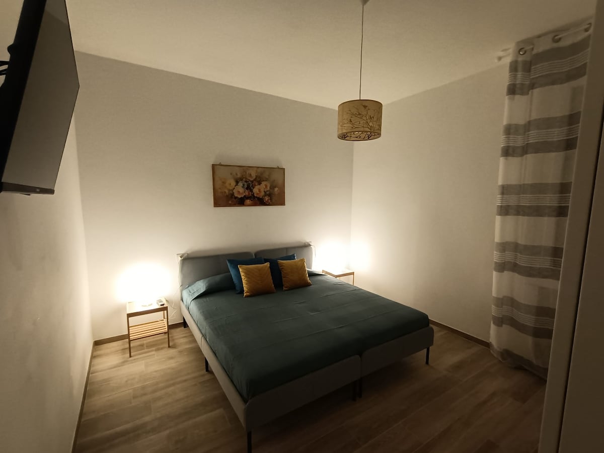 A serene sleeping area is presented with a spacious bed topped with a deep blue bedspread and accented by two yellow decorative pillows. Soft nightstands with lamps provide gentle illumination. A floral artwork adorns the wall above the bed, complemented by light curtains on the nearby window.