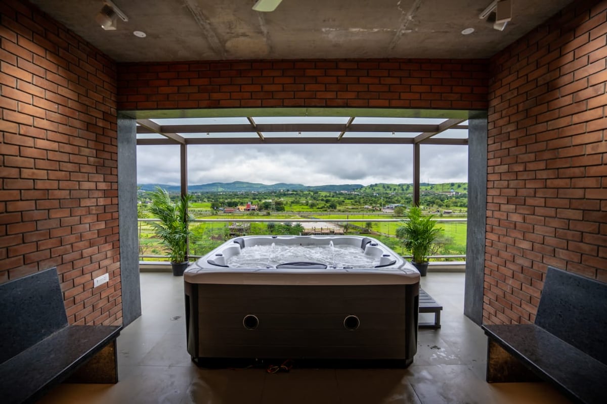 A spacious jacuzzi is situated against a backdrop of large glass doors, revealing expansive views of lush greenery and distant hills. The brick walls add a touch of warmth, while two benches are positioned nearby for relaxation.