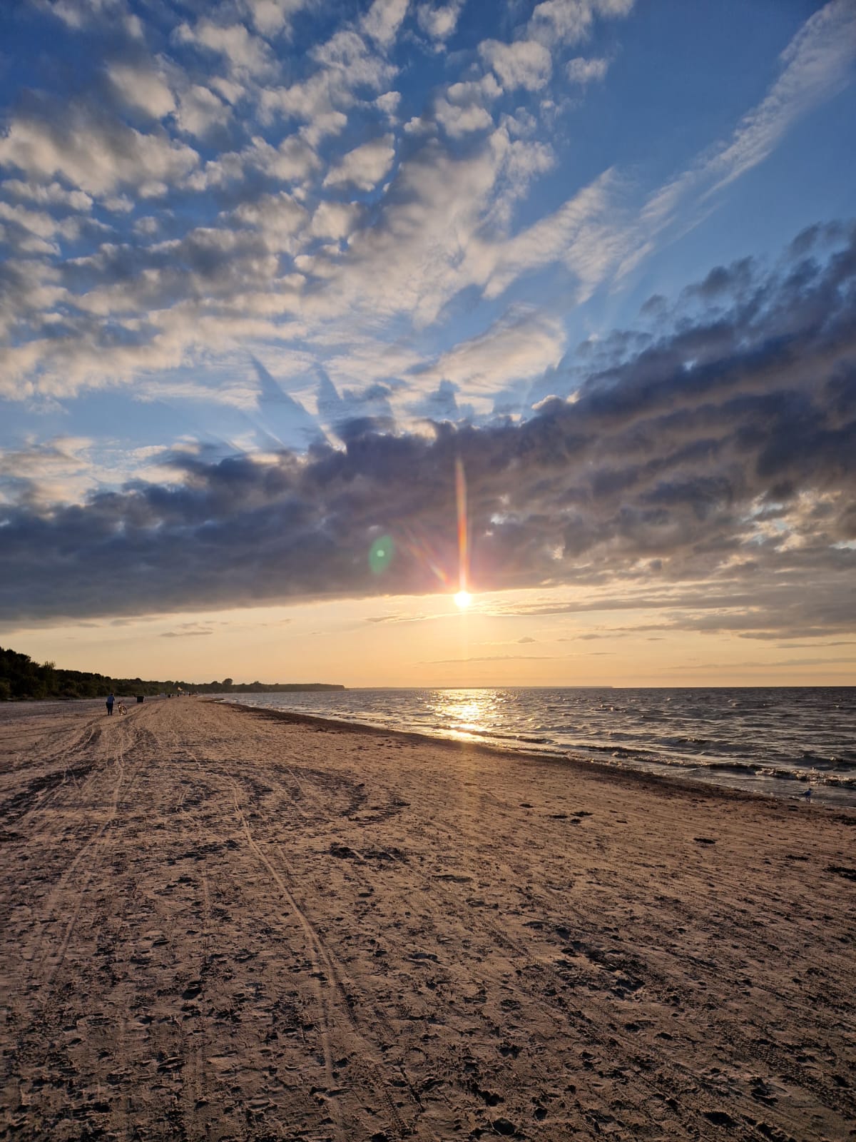 A serene beach scene is captured at sunset, with soft sand leading toward gentle waves. The horizon is adorned with a gradient of colors, reflecting the golden sun peeking through the clouds, while shadows stretch across the beach.