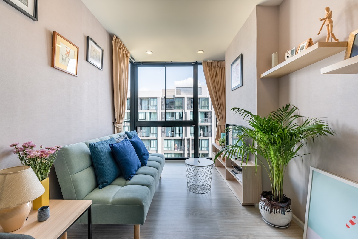 A cozy living area is visible, featuring a soft green sofa adorned with blue pillows. A small coffee table sits in front, with a decorative plant in one corner. Natural light floods the space through large windows, while light beige walls create a calming environment.