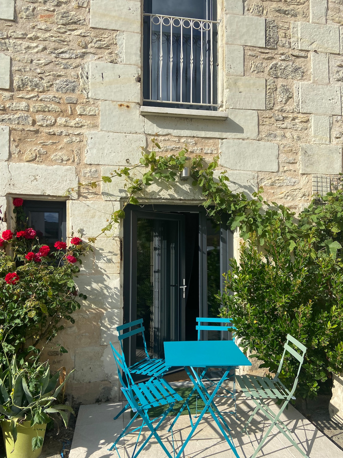 An outdoor seating area is set against a charming stone facade. A turquoise table is surrounded by four matching chairs, while vibrant flowers and greenery frame the entrance, enhancing the welcoming atmosphere.