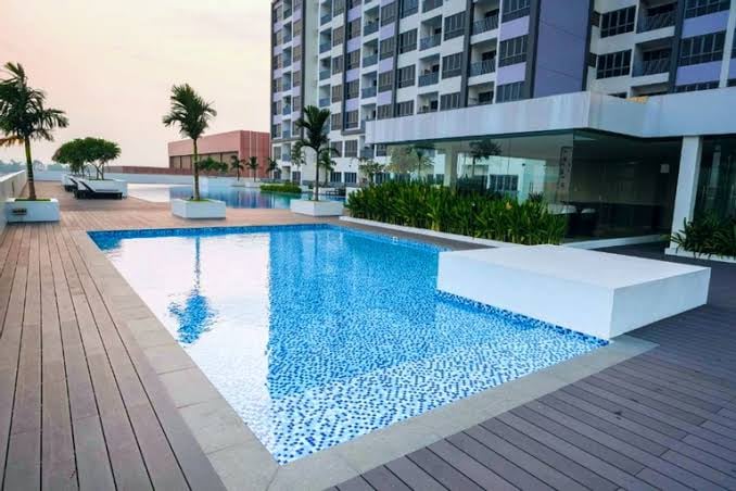 A modern outdoor pool is surrounded by beautifully arranged decking and tropical plants. Clear blue water reflects the sky, while lounge chairs are positioned nearby for relaxation. A sleek, contemporary building serves as a backdrop, enhancing the space's tranquil ambiance.
