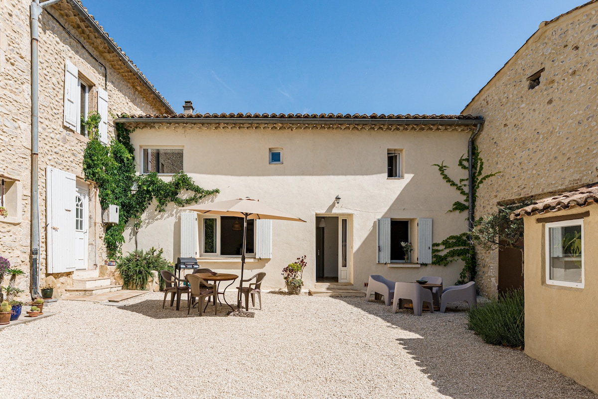 A charming courtyard features a light-colored stone facade adorned with climbing greenery. A large umbrella shades a dining table surrounded by chairs, offering a serene outdoor space. The gravel ground is neatly arranged, and the entrances to the surrounding buildings are visible, providing a welcoming atmosphere.