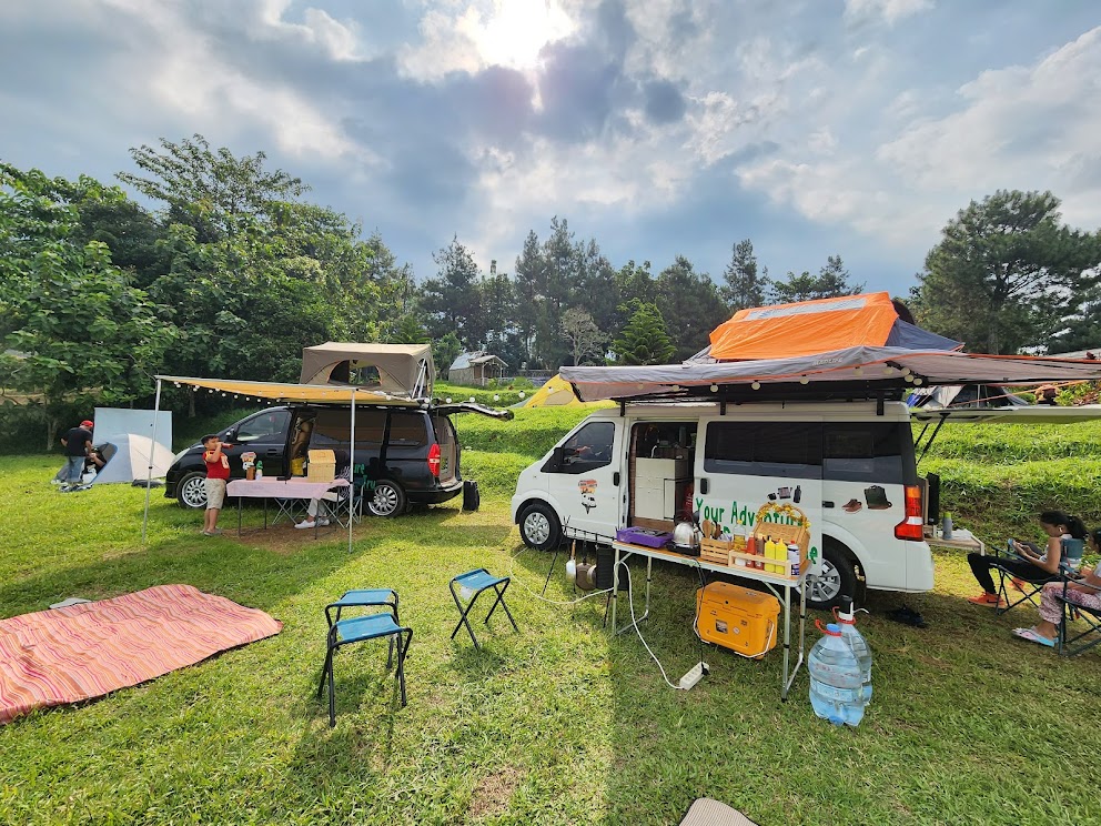 A spacious outdoor area is shown with two vans parked under a partially cloudy sky. Folding chairs and a table are set on the grass, while a colorful picnic blanket is laid out. Lush greenery surrounds the campsite, creating a natural atmosphere.