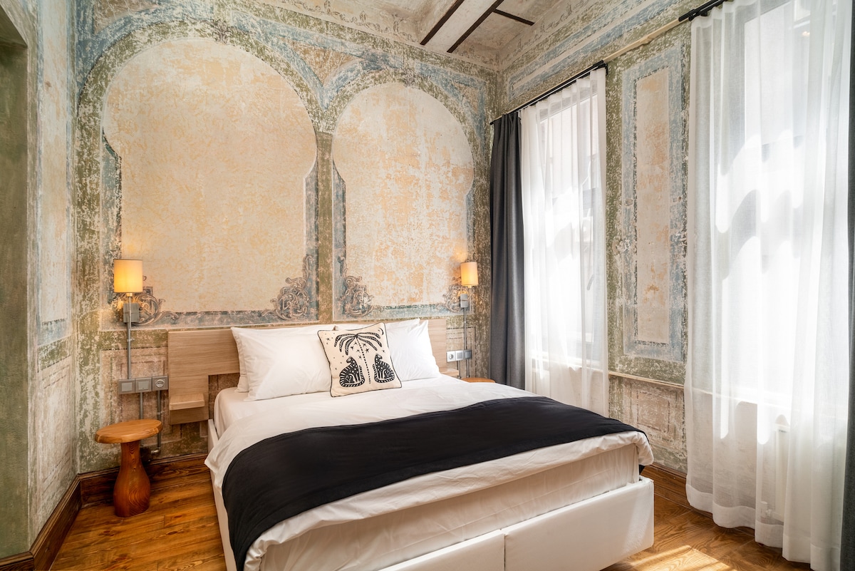 A spacious bedroom features an elegantly designed bed with a dark bedspread atop white linens. The walls are adorned with intricate decorative patterns, highlighted by natural light from tall windows dressed in sheer curtains. Soft lighting from bedside lamps contributes to a serene ambiance.