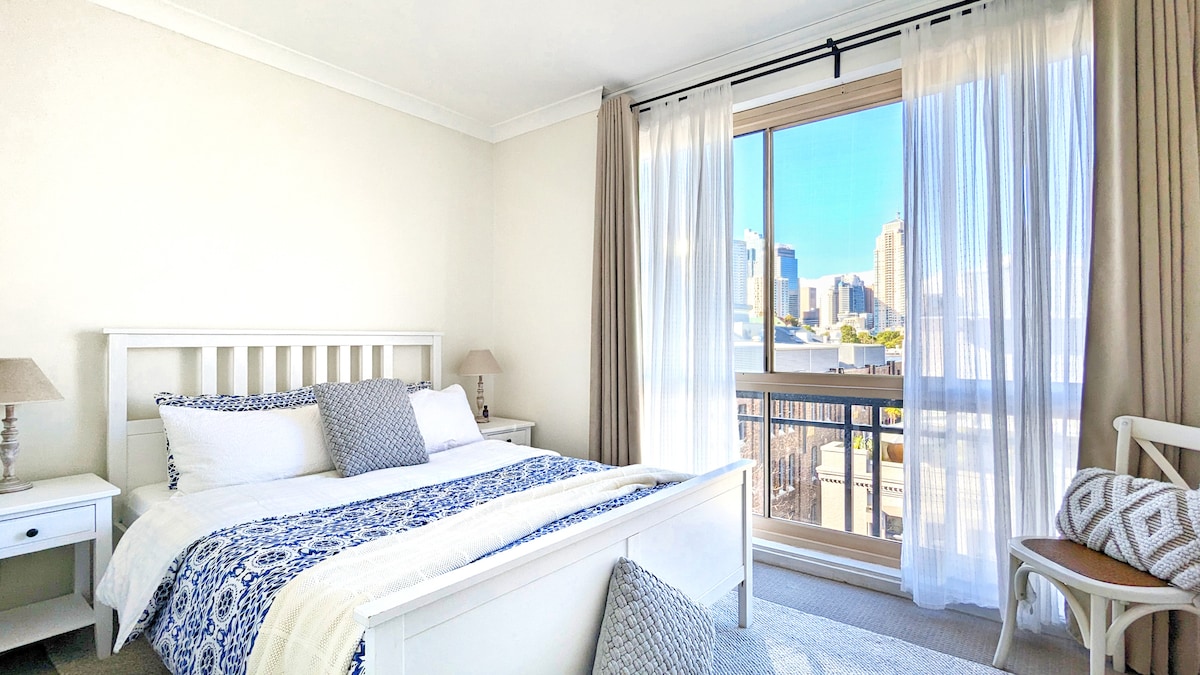 A bright bedroom features a spacious bed adorned with a blue and white patterned throw. Soft natural light filters through sheer curtains, illuminating the room. A small bedside table complements the calming décor, while a seating area offers a view of the city skyline through the balcony doors.