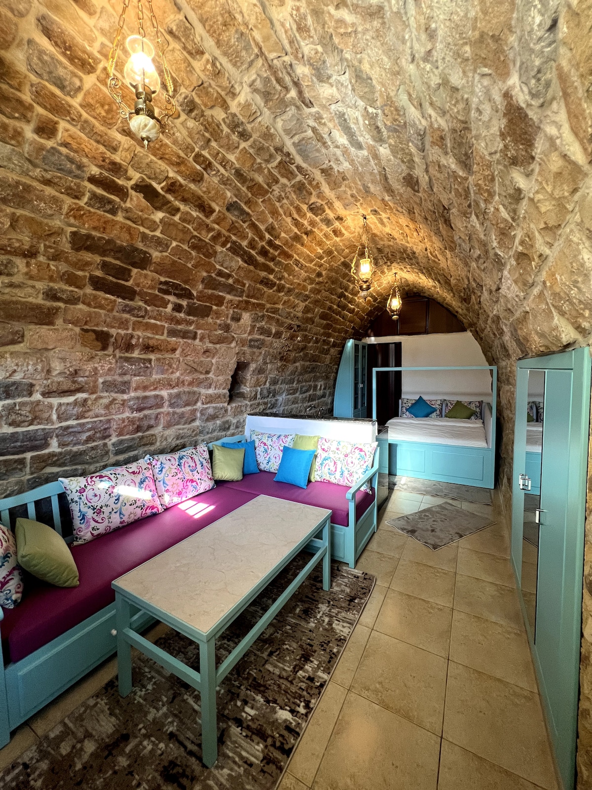 An arched stone ceiling enhances the cozy seating area, which features colorful floral-patterned cushions on a long bench and a light blue table at the center. Soft lighting from hanging fixtures creates a warm ambiance, while a nook with additional seating is visible in the background.