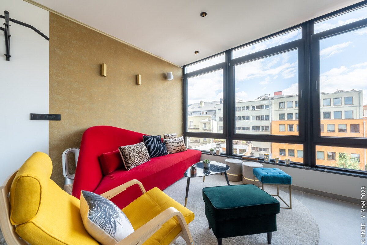 A spacious living area is highlighted by large windows that allow natural light to fill the space. A red sofa is paired with a mix of decorative cushions. Two accent chairs and a glass coffee table complete the arrangement, creating a comfortable environment.