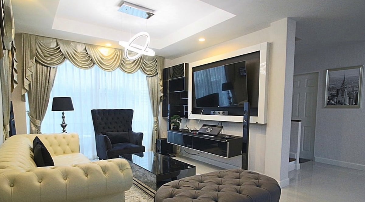 The lounge area features a cozy white sofa and a plush ottoman, complemented by a dark armchair. A large flat-screen television is mounted on a stylish wall unit. Natural light filters through sheer curtains, enhancing the modern decor accentuated by soft lighting fixtures.