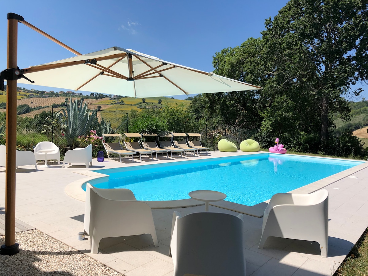 A modern outdoor space features a large swimming pool surrounded by sun loungers and white chairs. An umbrella provides shade over the seating area, with lush greenery and rolling hills visible in the background. A few colorful inflatables float in the pool.