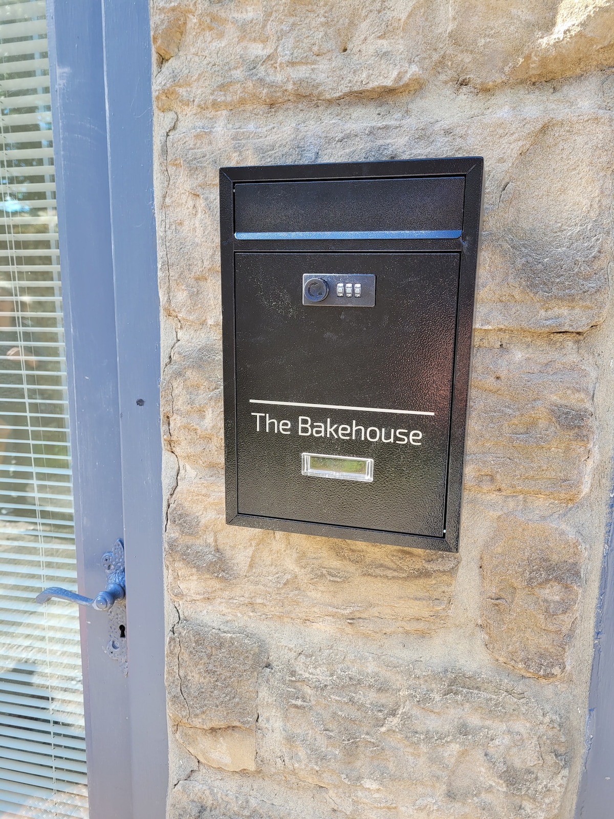 A black mailbox labeled 'The Bakehouse' is mounted on a stone wall. The mailbox features a small keypad and a slot for mail. Natural light enhances the texture of the stone, highlighting the rustic character of the building's exterior.