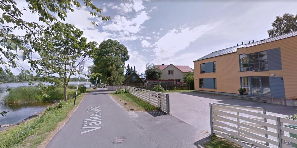 The image captures a serene street view featuring a calm lake on one side and a row of buildings on the other. A gravel driveway leads to a modern structure with mixed-color siding, framed by trees and landscaped greenery.
