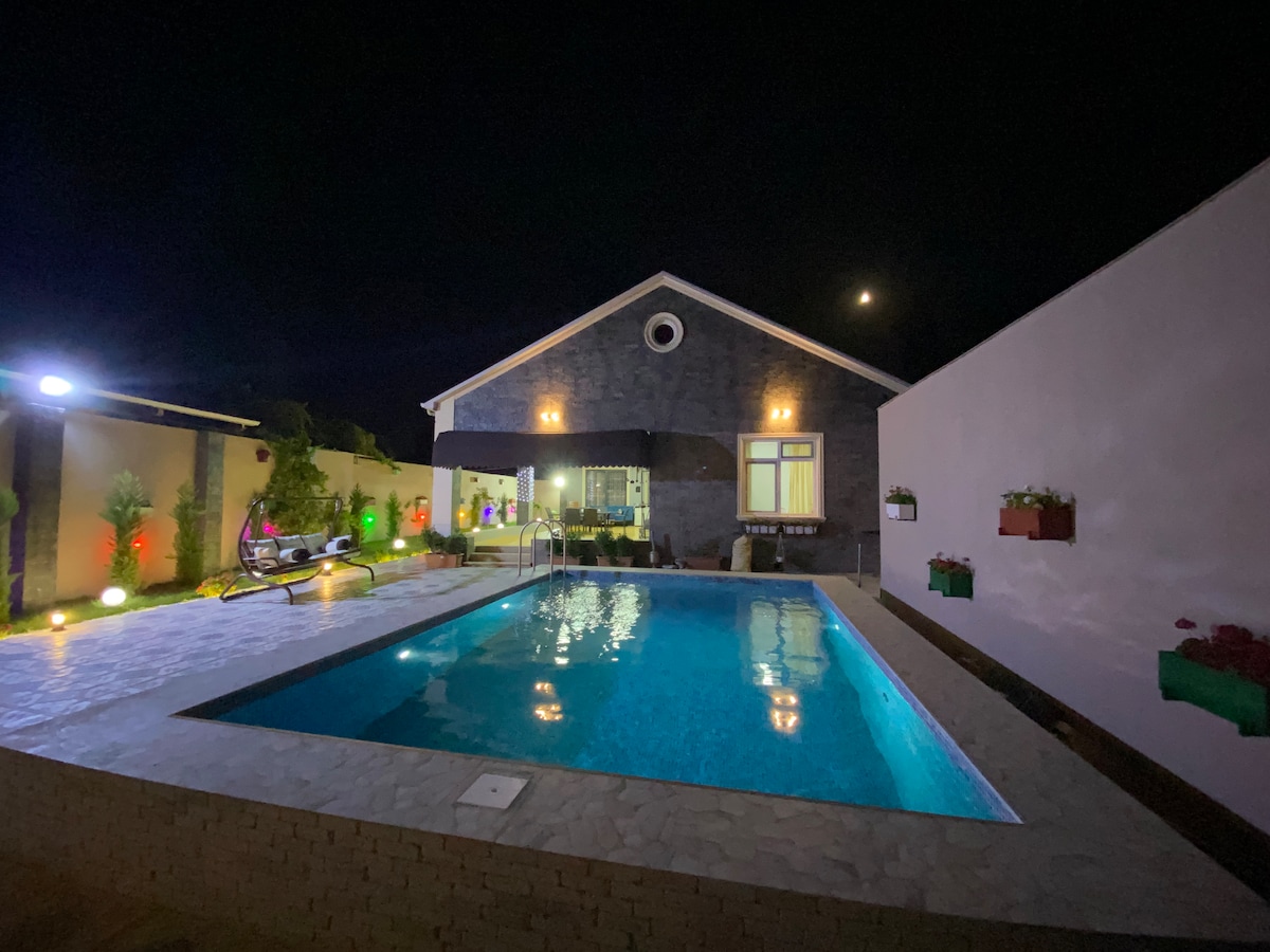 A modern villa is illuminated at night, featuring a clear swimming pool reflecting soft lights. The exterior is framed by walls adorned with vibrant planters. Panoramic windows allow for views of the inviting indoor space, creating a seamless connection with the outdoor area.