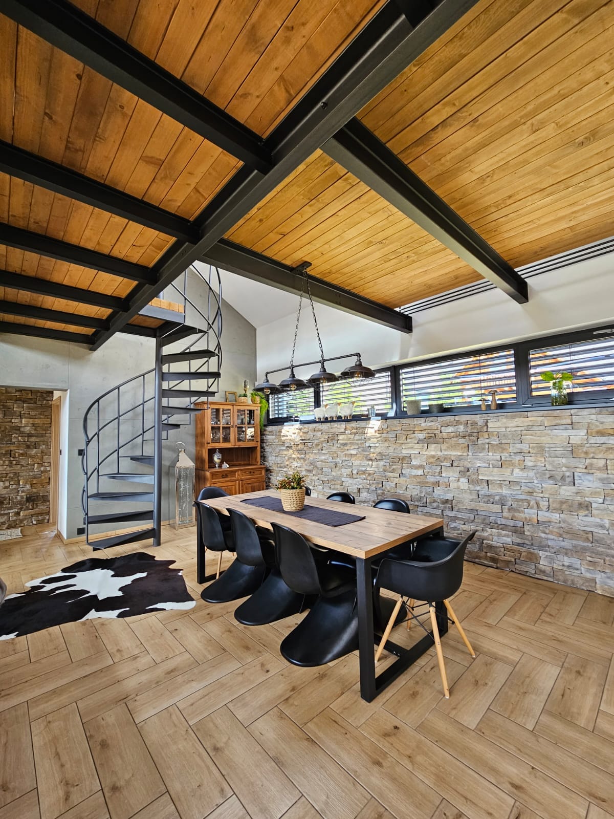 A modern dining area features a wooden table surrounded by black chairs, set against a textured stone wall. A staircase spirals up, complementing the open ceiling with wooden beams. Natural light filters through the windows, highlighting the earthy tones of the decor.