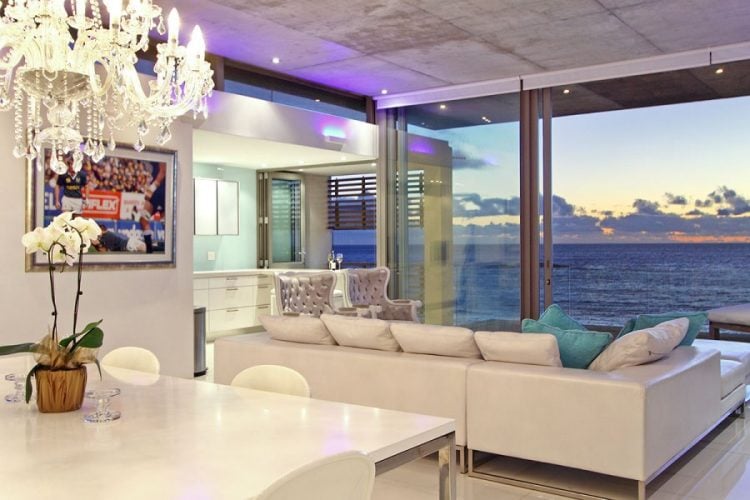 An open-plan living area features a modern design with large glass panels offering stunning ocean views. A chic chandelier hangs above a white dining table, while a light-colored sofa adorned with cushions invites relaxation. The space is illuminated by soft ambient lighting, enhancing the coastal vibe.