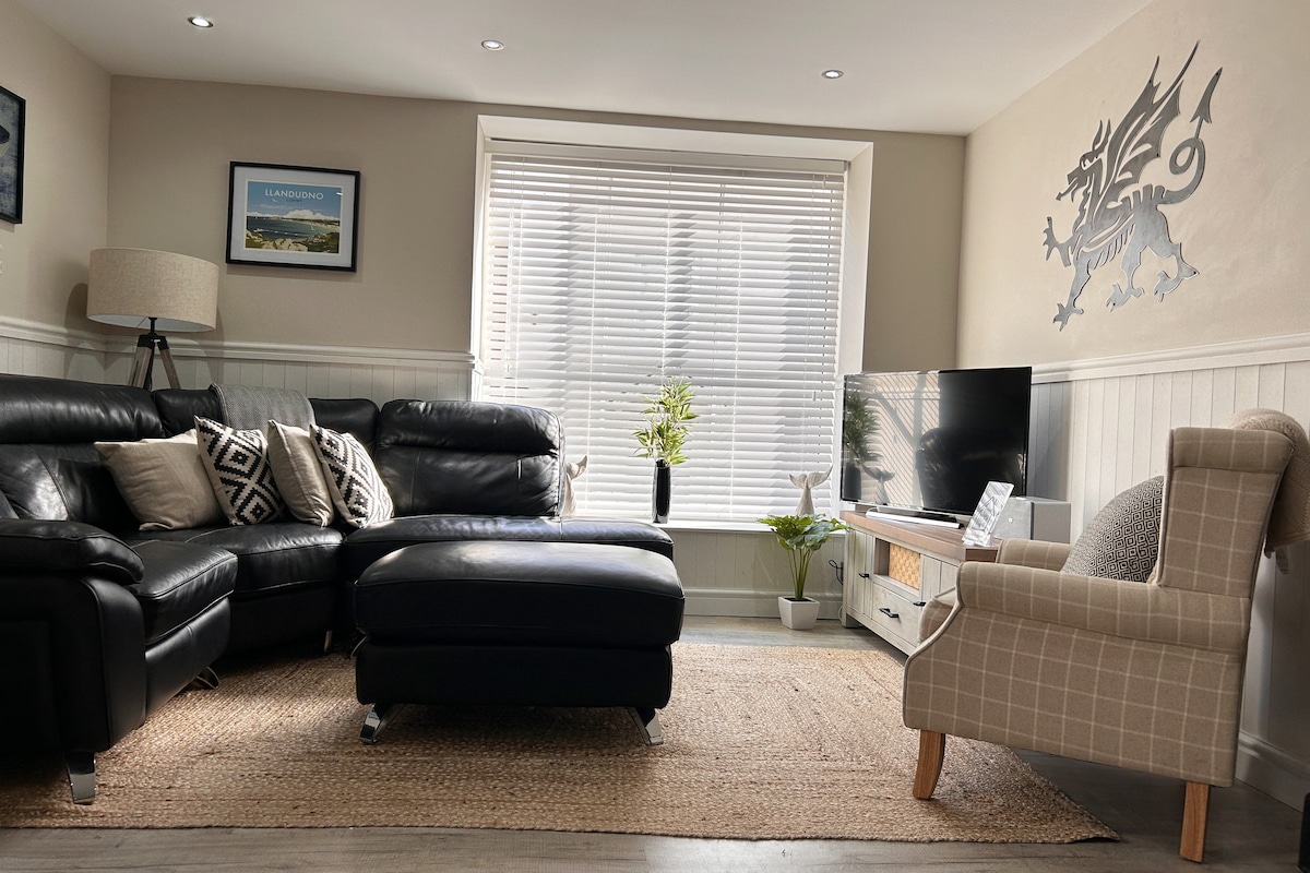 A comfortable living room features a large sectional leather sofa with decorative cushions arranged around a soft, woven rug. A flat-screen TV is positioned on a light wood unit, and natural light filters through the window, which is covered with horizontal blinds. A potted plant adds a touch of greenery.