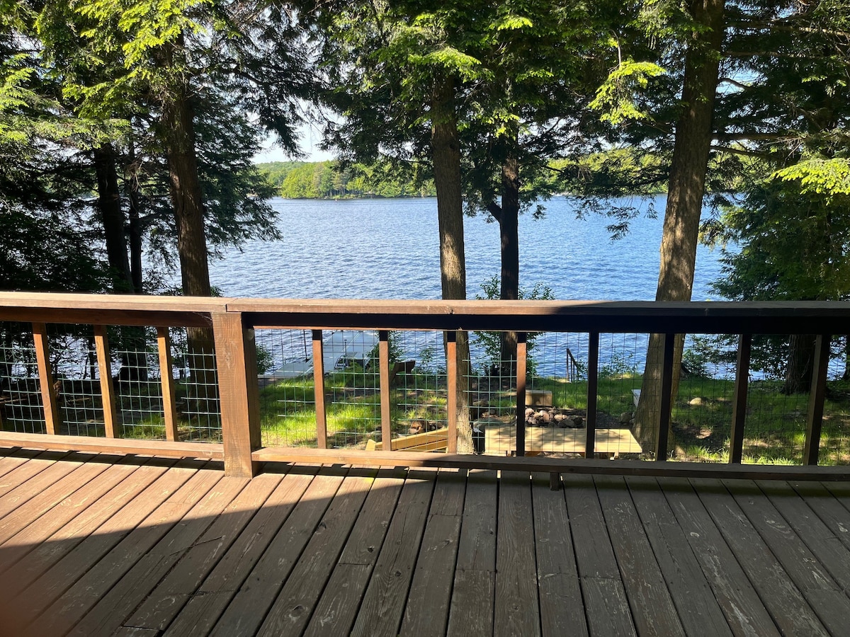 The outdoor deck provides a view of Cranberry Lake surrounded by tall trees. The calm water reflects the greenery, while a wooden staircase descends from the deck towards the water's edge, leading to a fire pit area.