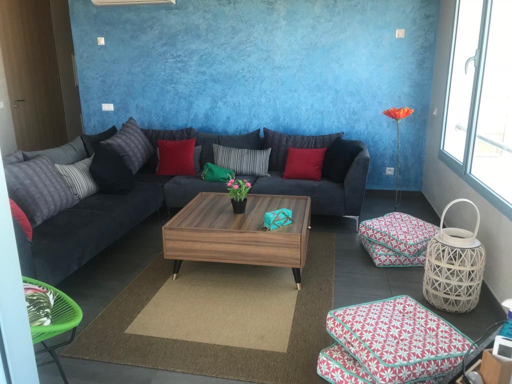 A modern living area features a dark blue sectional sofa adorned with an array of colorful cushions. A wooden coffee table is centered on a patterned rug, with decorative seating cushions placed around it. Sunlight filters through large windows, illuminating the space.
