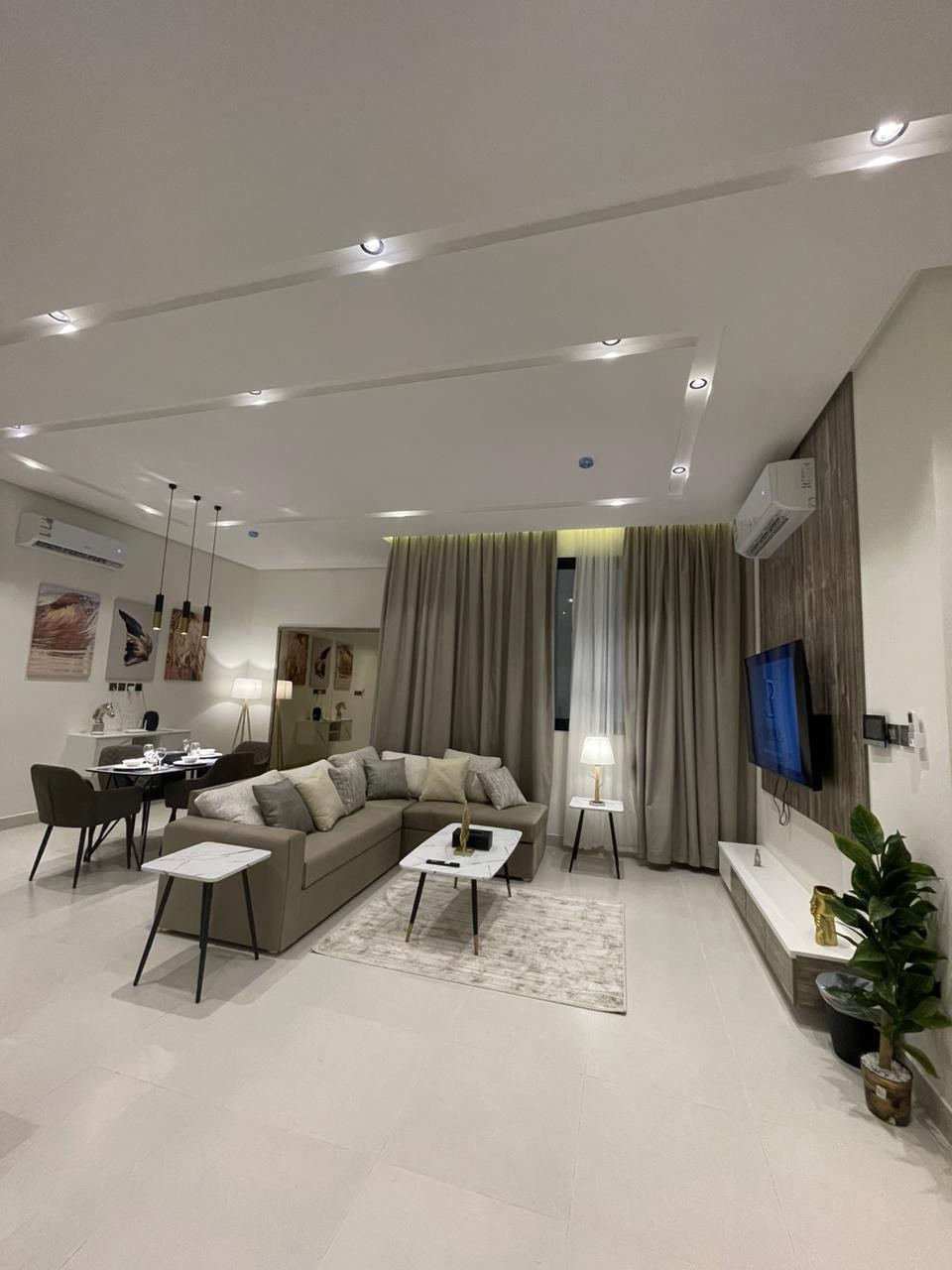 A modern living area features a spacious sectional sofa adorned with throw pillows. A rug lies beneath a coffee table. In the background, a dining table and chairs are visible. Warm light is provided by a table lamp next to the window, framed by long curtains.