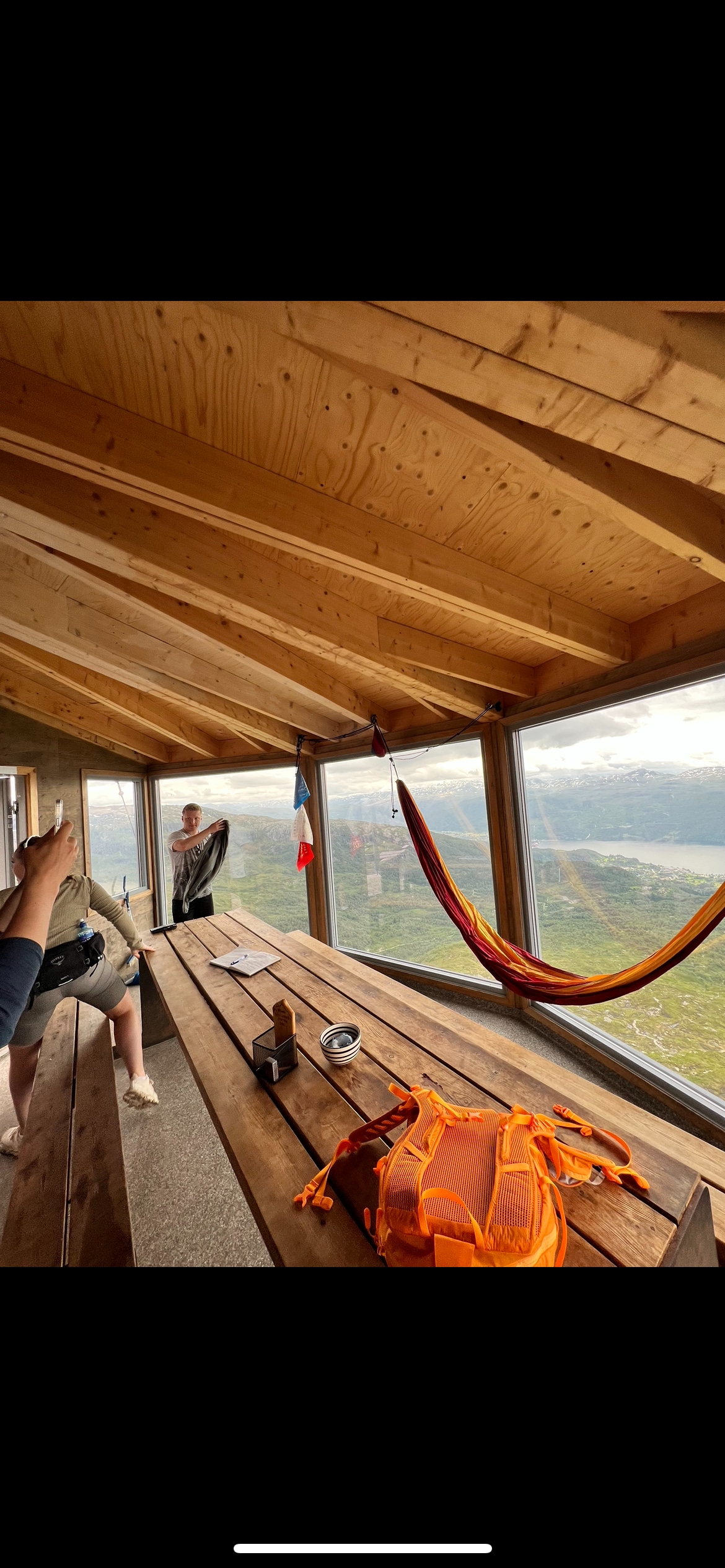 A spacious indoor area features a long wooden table surrounded by benches. Large windows offer expansive views of the landscape. A colorful hammock hangs from the ceiling, adding a relaxed ambiance to the space, while wooden beams create a warm and inviting ceiling.