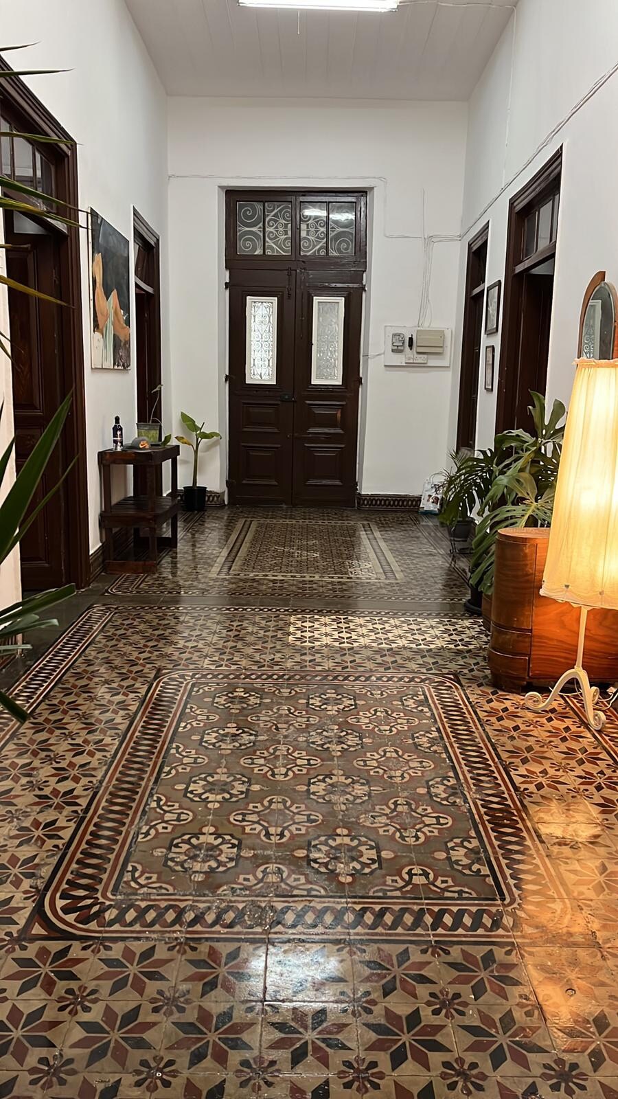 A wide hallway features a patterned tile floor with intricate designs. Tall, wooden doors are positioned at the end, allowing natural light to brighten the space. Potted plants are positioned along the walls, and a tall lamp stands on the right, adding warmth to the corridor.
