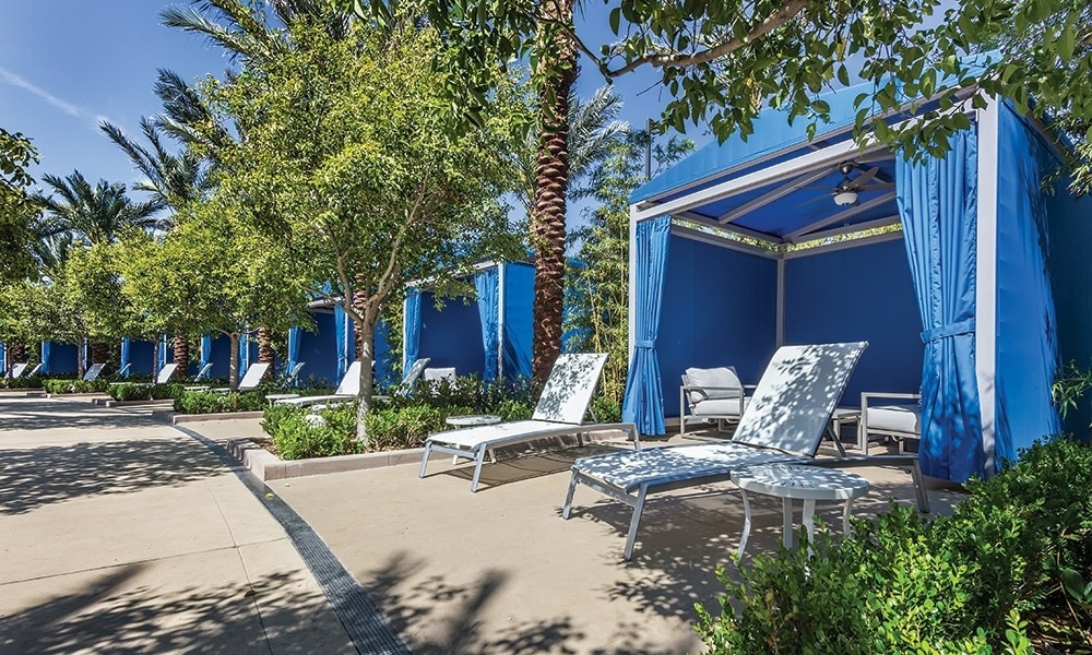 Lounge areas are surrounded by lush greenery and vibrant blue cabanas. White sun loungers are arranged under the shade of palm trees, offering a comfortable spot for relaxation. The bright blue design creates a refreshing atmosphere in this tranquil outdoor space.
